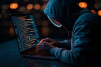 Hooded figure typing code on laptop in dark room with orange bokeh lights, representing cybersecurity threat, hacking or cybercrime concept