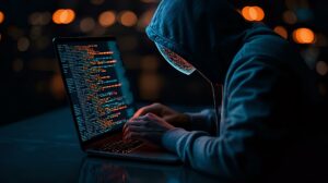 Hooded figure typing code on laptop in dark room with orange bokeh lights, representing cybersecurity threat, hacking or cybercrime concept