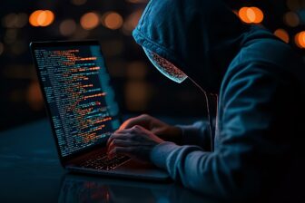 Hooded figure typing code on laptop in dark room with orange bokeh lights, representing cybersecurity threat, hacking or cybercrime concept